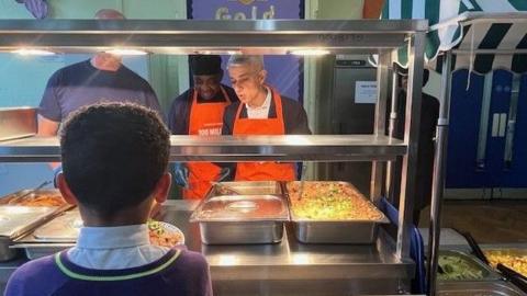 Sir Sadiq Khan is standing behind a glass hot food counter in a school as though he is serving food. He is standing alongside two staff members. He is looking down and appears to be talking.
A boy has his back the camera and faces the food counter as though waiting to be served.