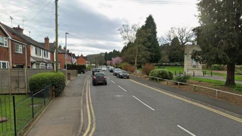 A Google street view of a single-carriage road in a residential area. Terraced houses are on the left of the road, while a park with a memorial is on the right. A number of cars are parked or driving on the road.