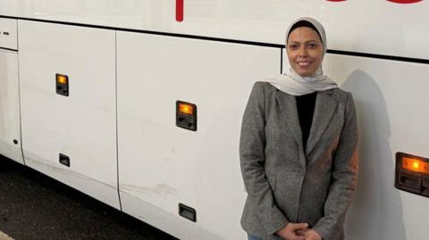 Fatima Benzbir wears a beige jacket, black top and white headscarf, as she leans casually against a coach.