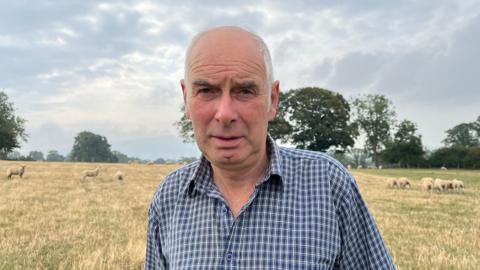 Photograph of Price, who is wearing a blue and white checked shirt, stood in a field with a serious expression. Behind him are about 10-15 white sheep. The field is covered in yellow-green grass, with trees lining the edge of the field. The sky is cloudy and overcast. 