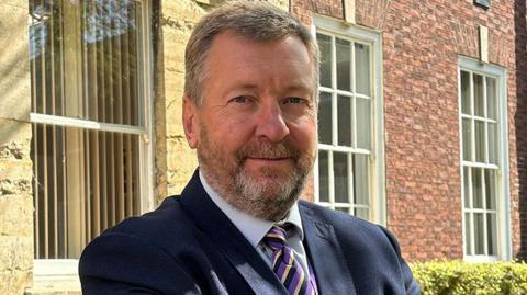 A man with blue eyes and short cropped grey-blond hair, with the same coloured beard and eyebrows. He is wearing a black, purple and cream striped tie from the collar of his blue shirt, over which is a navy blazer. He is standing in front of a red and stone-coloured brick building with Georgian-style windows.