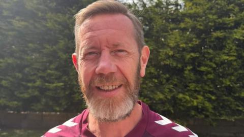 Colin Brown has short fair hair and smiles at the camera. He wears a maroon Hearts FC strip and is standing in a garden with a bush behind him