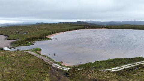 Aerial image showing a loch with a small beach along the furthest shore and vehicle track passing the closest edge. 