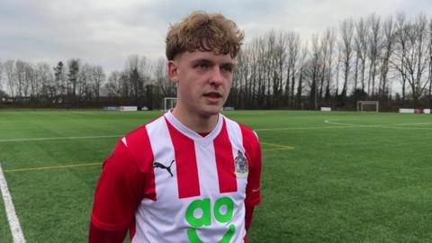 A young man with blonde, short curly hair, wears an Altrincham football shirt, standing on a football pitch 