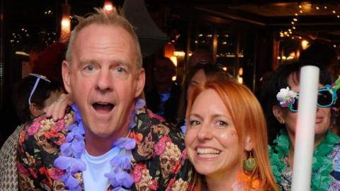 The DJ Fatboy Slim, wearing a floral short-sleeve shirt over a white t-shirt with a garland of flowers around his neck, stands beside a woman with red hair and in front of two people also dressed in bright clothing and wearing sunglasses.