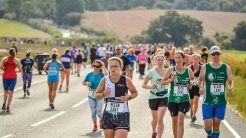 A large group of runners jogging along a road with green fields either side and trees in the distance.