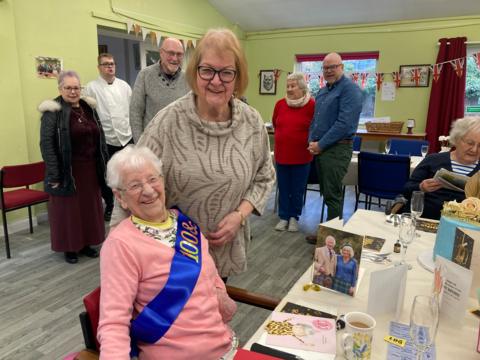 Lady smiling whilst sat on a red chair at a table with birthday cards and a cup of tea and champagne glasses and birthday cake. She is wearing a pink jumper with a dark blue sash with gold writing that says 100 and fabulous. A lady is stood behind her and is smiling and wearing a beige jumper. A group ofpeople are stood behind them both.