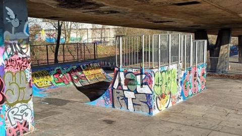 A skate park sits underneath a concrete bridge. A concrete ramp, covered in graffiti sits in the middle of the concrete floor. 