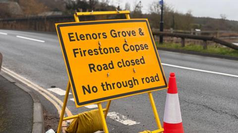 Image shows a road with traffic. There is a sign and a traffic cone.
A yellow information sign saying that Elenors Grove & Firestone Copse Road closed, No through road.