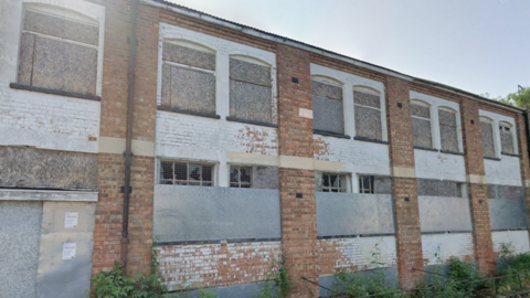 The facade of an old brick built factory, whose windows are boarded up or broken. It is two storeys and the bricks are a mix of white and sand coloured. Wild shrubs grow along the front