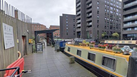 A number of barges are situated along the marina. On the left is the wooden outbuilding while on the right is a number of flat blocks. 