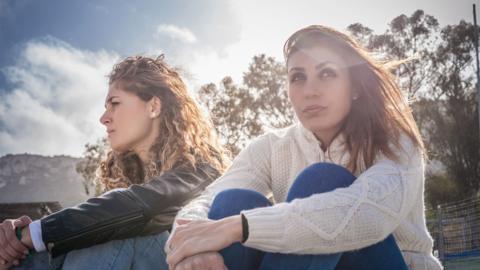 
Two sullen young women friends sitting on breezy beach - stock photo