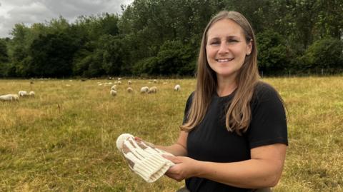 A woman stood in a field. She has long brown hair. She is wearing a black top and holding a woolly hat. Behind her there are sheep in the distance.