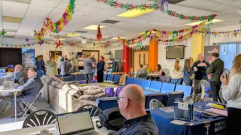 The picture shows a busy community event taking place in a large indoor hall decorated with colourful festive garlands and bunting. There are several tables set up around the room, some with laptops and paperwork, suggesting advice or support services are being offered. People are seated at tables and others are standing in small groups, talking and engaging with each other.