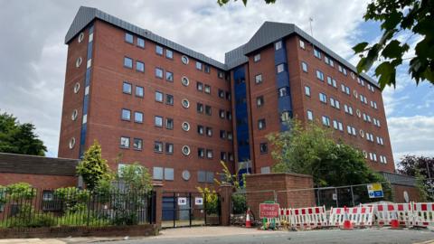 Two large tower blocks made of red bricks, blue cladding and grey panels stand behind a metal fence. The lower doors and windows are covered with metal sheeting. A sign next to the gate reads 'Road closed'.
