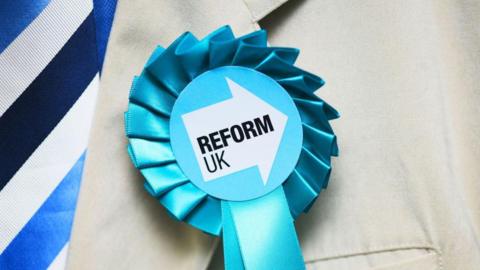 A Reform UK rosette is attached the the lapel of a beige coloured jacket. The rosette is pale blue. In the centre it has a white arrow pointing to the right with "Reform UK" in black ink. 