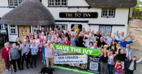 About 30 supporters - standing outside the Ivy Inn, which is a traditional old pub with whitewashed walls and thatched roof - all with their thumbs up in the air and smiling. They are holding a large banner saying 'Save the Ivy'.
