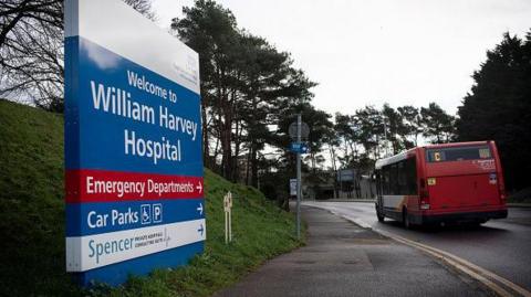 The entrance to William Harvey Hospital in Ashford. A large blue, white and red hospital sign in the foreground reads "Welcome to William Harvey Hospital" with directions to the emergency department, car parks and the Spencer private consultants unit. A red bus is driving away along the road beside the hospital grounds, with trees and hospital buildings visible in the background.