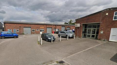 An industrial estate with a few parked cars and steel shutters drawn down, on an overcast day.