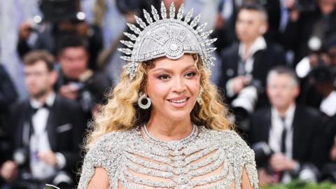 US performer Beyonce poses on the red carpet for the 2026 Met Gala wearing a shimmering skeletal gown and a spherical headpiece as photographers in suits stand behind her.