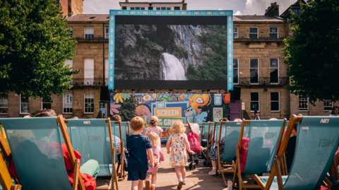 A large screen displaying a picture of a waterfall stands in front a row of buildings in Newcastle's Eldon Square. Pointed at the screen are dozens of blue deckchairs where people are sitting to watch the film. Five children are running between the chairs towards the screen.