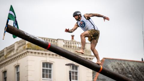 A man in shorts and a singlet races up a mast to capture a Devon flag.