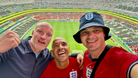 Ashley Price has his arm around Stan Collymore who is wearing a red football top with the England crest on his chest. A third man is wearing a blue England hat, and also has on a red England top. 