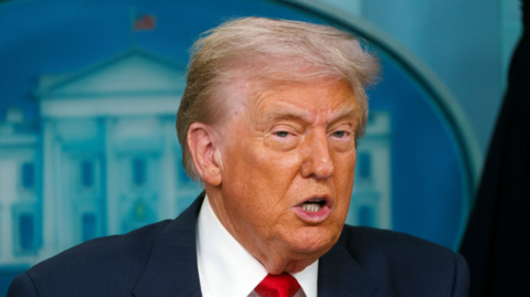 Close up of Donald Trump mid-speech, he's wearing a dark blue suit, white shirt and red tie, the White House logo affixed to the blue wall behind him