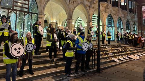 Lots of protestors in high visibility jackets stand outside the Guildhall council building holding circular face signs depicting the impact of unclean air in Northampton.