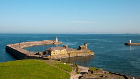 A sunny day looking over to Whitehaven harbour. There is green grass and the sea is very calm. 