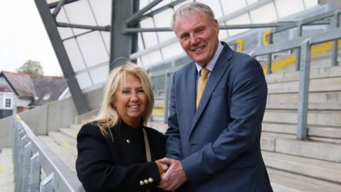 Gill Spencer stands close to Andrew Thirkill in the stand of a stadium. The pair are locked in a handshake and smiling into the camera. Gill is wearing a smart black coat with brass buttons while Andrew is in a blue suit and yellow tie.