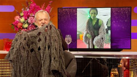 Graham Norton holding up a tattered cardigan, in the background there is a television screen showing Cynthia Erivo as Elphaba in Wicked: For Good, wearing the cardigan