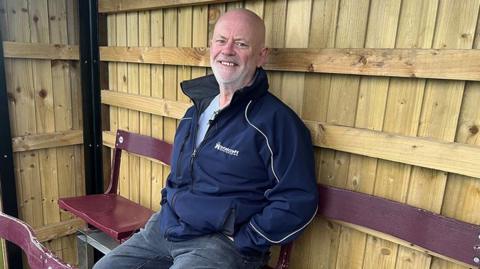 Ian Nutter sits in the timber clad stand. He is sitting on a burgundy seat and it wearing jeans and a blue zip up casual jacket. He is bald with a short white beard and is smiling.