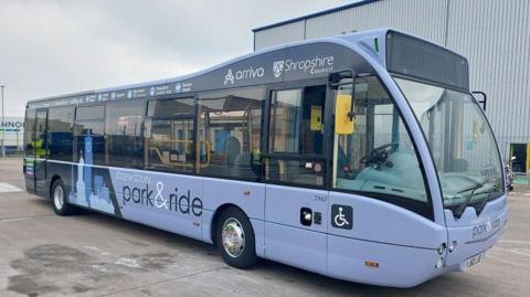 A lilac coloured park and ride bus. It has Shropshire Council and Arriva logos on it, with a decal of the silhouette of a clock tower and other buildings.