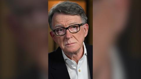 Peter Mandelson is wearing black glasses, a white unbuttoned shirt and a black blazer. He is pictured while being interviewed by the BBC. The background is orange and out of focus.