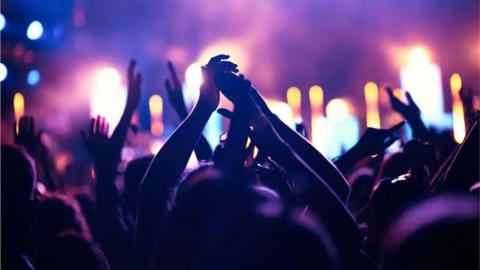A night club, where several people are seen in silhouette raising their arms whilst dancing. Lights are visible in the background.