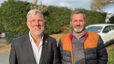 Deputy Adrian Gabriel on the left - A man with white hair wearing a black suit on top of a white shirt. He has a Guernsey lapel pin on his left lapel. On his right is Deputy Marc Leadbeater - a man with black and grey hair, wearing an orange and black puffer jacket, on top of a grey fleece and a grey T-shirt. 