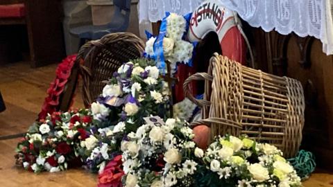 Two wicker baskets lean in either side of wreaths of flowers and a lifeboat ring.