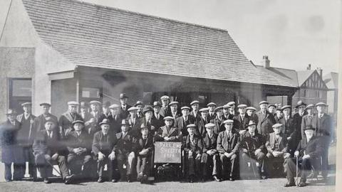 A black and white photograph of a large group of older men, all wearing headgear of one sort or another, outside the Park End Shelter. The man in the centre holds a sign which dates the picture as 1938. The shelter is a single-storey building with a very tall roof which overhangs in the front to provide a covered area.