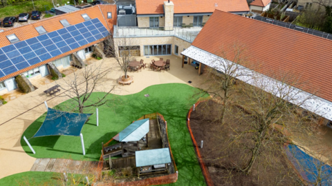 A birds eye view of a large building wrapping around outdoor space including tables and chairs, trees, greenery and a children's play area.