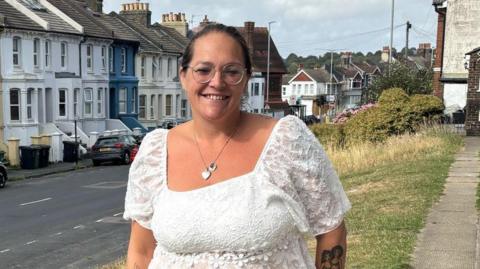 Kelly Warwick standing outside her house in Brighton. She wears a white blouse and dark jeans. She has dark hair tied up, and glasses.