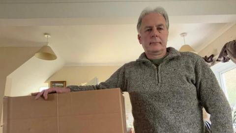 Marcus Kats. He is wearing a grey jumper and has his arm outstretched on a cardboard box.