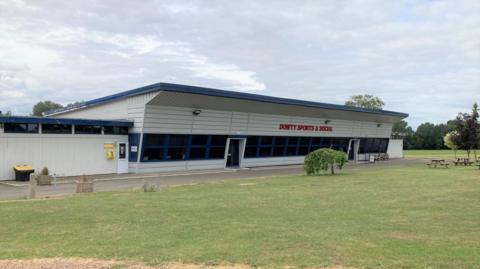 A large grey building with a slanting roof. The words "Dowty Sports & Social" are written in large red letters above the entrance. Grass surrounds the building and there are several picnic benches and trees in the distance.