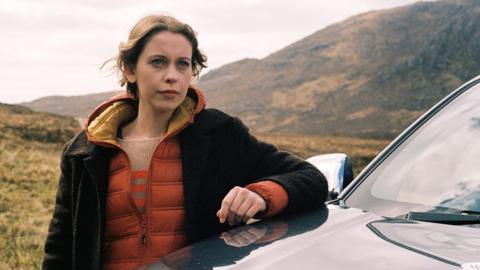 Star Sorcha Groundsell as Kat in An t-Eilean (The Island) - she is outside leaning on a car bonnet, with the hills pictured behind her. She has auburn hair, a pensive look on her face and a brown and orange jacket on.