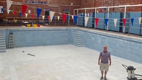 Brian McCann wearing a purple and white striped T-shirt and cream shorts stands smiling in the middle of the empty pool Pacific Pool with a smile on his face. Hanging above him and the pool are multi-coloured backstroke flags. There are tools and equiptment on the side of the pool. 