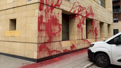 Red paint has been sprayed across the yellow bricks of the building. It has dripped down the wall and dyed the pavement red.
