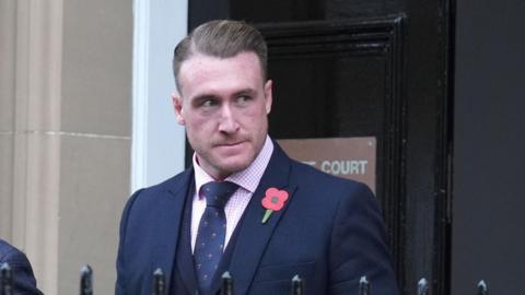 Stuart Hogg grimacing as he comes out of court. He is wearing a suit and tie with a poppy attached to his jacket.