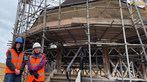Emma Hampton on the left is looking directly at the camera and smiling. She is wearing a black jacket with an orange fluorescent hi-vis vest over the top and blue hard hat. Next to her is Sarah Morrison, a woman who is also looking directly at the camera and she is wearing a purple outdoor jacket, orange fluorescent hi-vis vest and white hard hat. Behind them is a bandstand covered in scaffolding while restoration works are carried out.