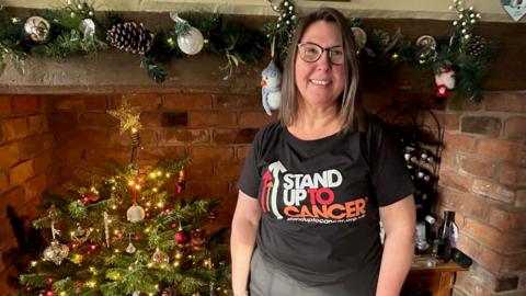 A woman with brown hair and glasses stood in front of a brick fireplace containing a Christmas tree. She is wearing blue jeans and a black t-shirt with stand up to cancer on it