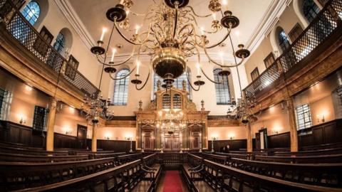 Interior of Bevis Marks Synagogue, featuring large gold candelabra-style chandeliers, dark wood seating and light-coloured walls